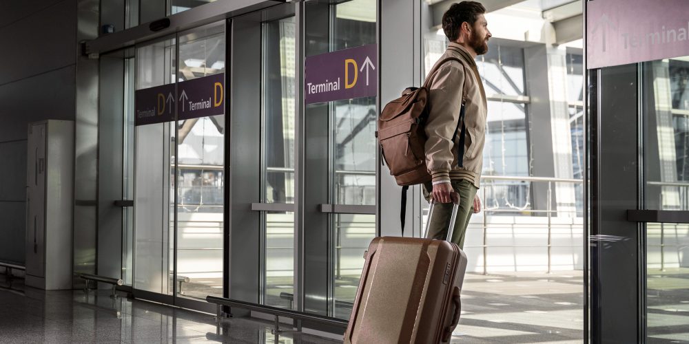 Bărbat cu rucsac și troler care iese din terminalul unui aeroport pentru a prelua o mașină închiriată.
