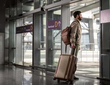 Bărbat cu rucsac și troler care iese din terminalul unui aeroport pentru a prelua o mașină închiriată.